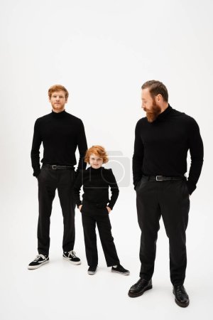 full length of bearded man looking at redhead son and grandson posing with hands in pockets on grey background