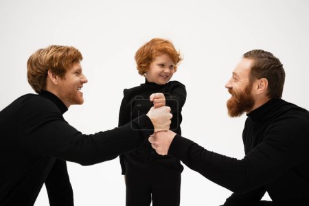 cheerful red haired kid with dad and bearded grandpa in black turtlenecks joining fists isolated on grey