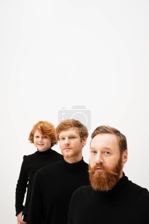 bearded man looking at camera near redhead son and smiling grandson in black turtlenecks isolated on grey