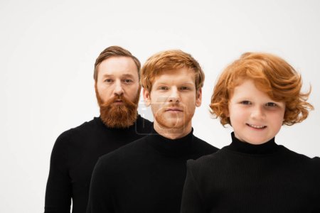 bearded man in black turtleneck looking at camera near redhead son and happy grandson isolated on grey