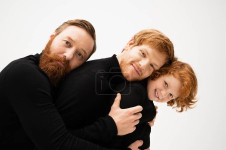 happy bearded man hugging redhead son and grandson in black sweaters isolated on grey
