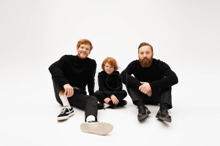 Photo for Full length of redhead men in black clothes posing near cheerful boy sitting with crossed legs on light grey background - Royalty Free Image
