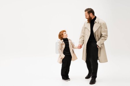 Photo for Happy redhead kid and bearded man in trench coats holding hands and looking at each other on light grey background - Royalty Free Image