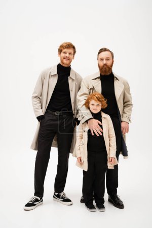 Photo for Full length of smiling red haired boy with father and grandpa in trench coats looking at camera on light grey background - Royalty Free Image