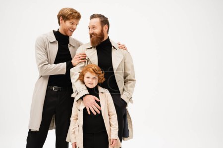 happy young man in trench coat hugging bearded father near redhead son isolated on grey