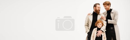Photo for Young man in trench coat hugging bearded father near redhead son looking at camera isolated on grey, banner - Royalty Free Image
