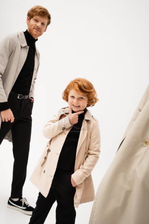 Photo for Cheerful red haired boy in trench coat touching chin while posing with hand in pocket near smiling dad on light grey background - Royalty Free Image