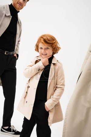Photo for Happy redhead boy in trench coat touching chin while posing with hand in pocket on light grey background - Royalty Free Image