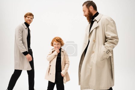 cheerful redhead boy touching chin while posing with hand in pocket near dad and grandpa isolated on grey