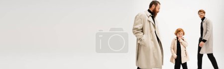 smiling redhead kid in trench coat posing with hand in pocket near father and granddad on light grey background, banner