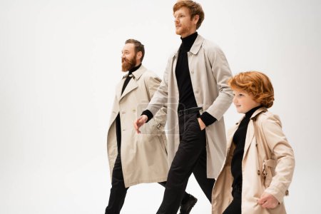Photo for Smiling red haired men and boy in stylish trench coats walking isolated on grey - Royalty Free Image