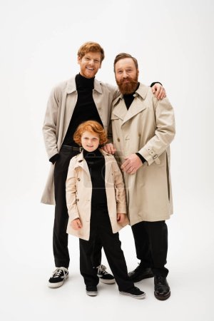 Photo for Cheerful man hugging bearded dad near redhead son while posing in trench coats on light grey background - Royalty Free Image