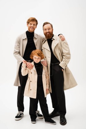 Photo for Joyful red haired man embracing bearded father and son standing in trench coats on light grey background - Royalty Free Image