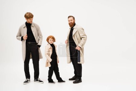 Photo for Red haired boy smiling at camera while standing with hand in pocket near dad and bearded grandpa in trench coats on light grey background - Royalty Free Image