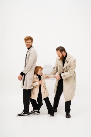 Photo for Full length of redhead man in trench coat smiling near bearded grandpa and grandson having fun on light grey background - Royalty Free Image