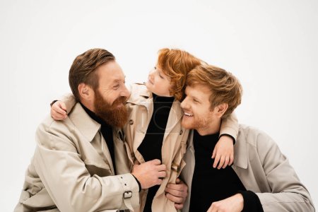 redhead kid embracing happy dad and grandfather smiling at each other isolated on grey