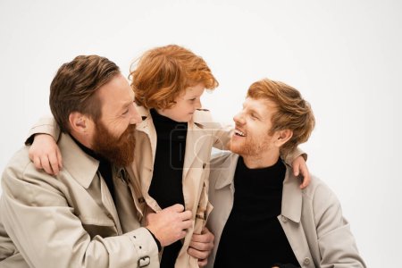 bearded man smiling near cheerful son and grandson in trench coats looking at each other isolated on grey