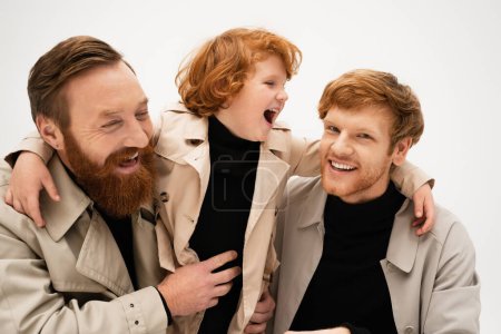 excited boy laughing and embracing father and bearded grandpa isolated on grey