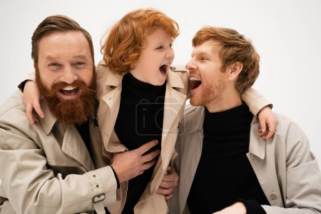 joyful bearded men and red haired boy in trench coats laughing isolated on grey