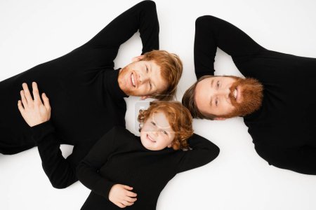 Photo for Top view of happy redhead men and boy in black sweaters looking at camera while lying on white background - Royalty Free Image