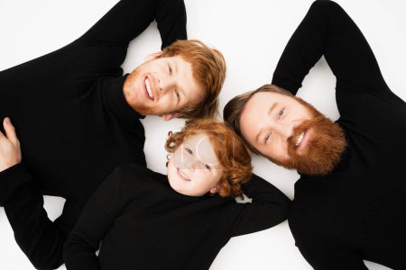 top view of smiling redhead boy with bearded grandfather and dad in black turtlenecks lying on white background