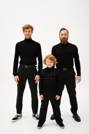 Photo for Full length of smiling boy and redhead bearded men in black turtlenecks and trousers standing on light grey background - Royalty Free Image
