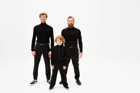 Photo for Full length of redhead men and kid in black sweaters and trousers standing and looking at camera on light grey background - Royalty Free Image