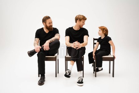 Photo for Redhead father and son looking at each other near smiling bearded man while sitting on chairs on grey background - Royalty Free Image