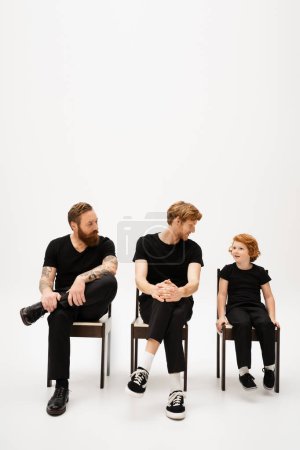 Photo for Bearded men in black clothes looking at smiling red haired boy while sitting on chairs on grey background - Royalty Free Image