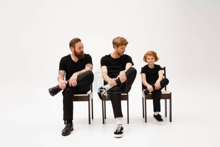 Photo for Red haired kid looking at camera near father and tattooed grandpa sitting on chairs in black clothes on grey background - Royalty Free Image