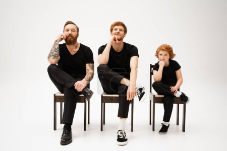 bearded men with redhead kid holding hands near face while sitting on chairs on grey background