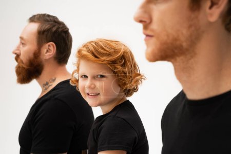 happy red haired boy smiling at camera near bearded tattooed grandpa and dad on blurred foreground isolated on grey