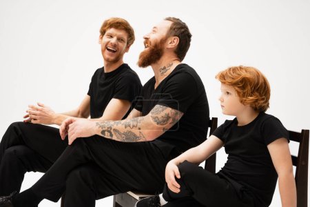 redhead boy sitting near tattooed grandpa and dad laughing isolated on grey