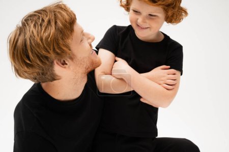 cheerful boy with crossed arms looking at redhead father isolated on grey