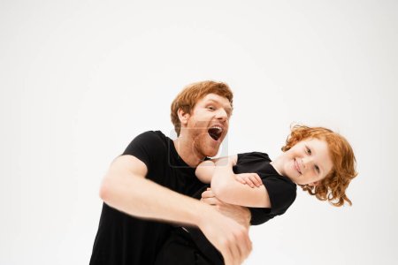Photo for Excited man playing with redhead son crossing arms and smiling at camera isolated on grey - Royalty Free Image