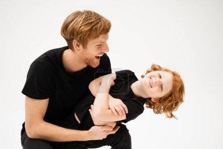 Téléchargez les photos : Excited man in black t-shirt playing with happy redhead son crossing arms isolated on grey - en image libre de droit