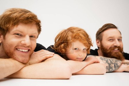 joyful man with redhead son and bearded tattooed dad lying with crossed arms and smiling at camera on light grey background