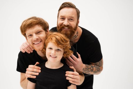 family portrait of redhead men and cheerful boy in black t-shirts hugging and smiling at camera isolated on grey