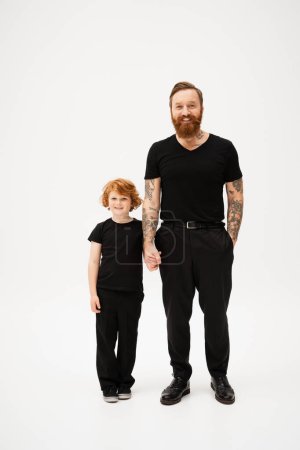 Téléchargez les photos : Full length of bearded tattooed man with redhead grandson holding hands and smiling at camera on light grey background - en image libre de droit