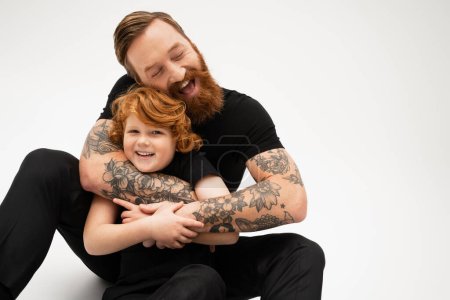 redhead boy smiling at camera near bearded and tattooed grandpa embracing him on grey background