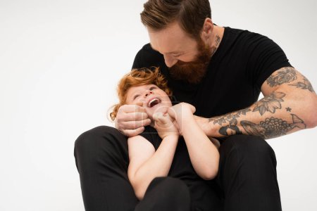 bearded and tattooed grandpa playing with excited redhead grandson isolated on grey