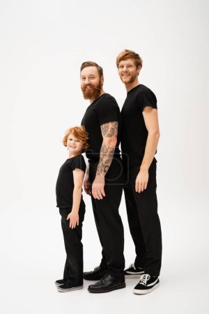 Photo for Full length of bearded redhead men and kid in black t-shirts and trousers smiling at camera while standing on light grey background - Royalty Free Image