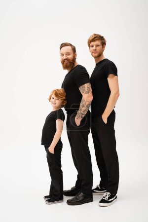 Photo for Full length of bearded tattooed man with redhead son and grandson posing with hands in pockets of black trousers on light grey background - Royalty Free Image