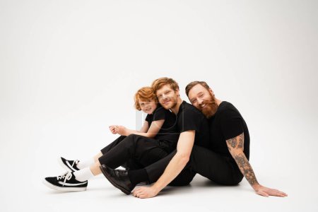 joyful redhead men and boy in black clothes looking at camera while sitting on light grey background