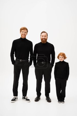 Téléchargez les photos : Full length of cheerful bearded men with red haired kid in black clothes standing with hands in pockets on light grey background - en image libre de droit