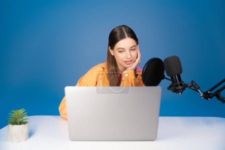 Teen podcaster looking at laptop near microphone isolated on blue 