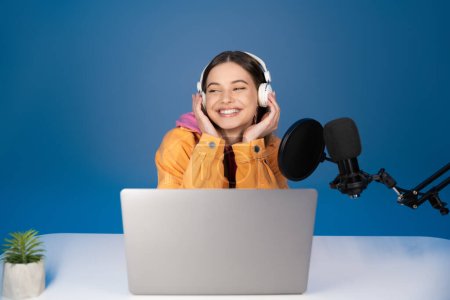 Cheerful teenager in headphones sitting near laptop and microphone isolated on blue 