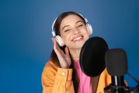Smiling teen girl in headphones looking at camera near blurred microphone isolated on blue 