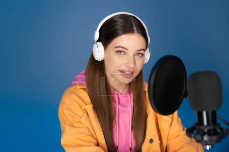 Téléchargez les photos : Smiling teen girl in headphones looking at camera near studio microphone isolated on blue - en image libre de droit