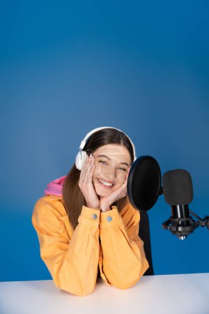 Smiling teenager in headphones sitting near studio microphone isolated on blue 
